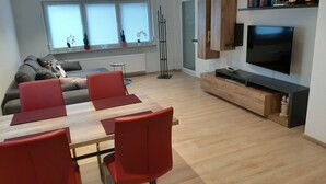 A modern living room with a sofa, red chairs, a wooden dining table, and a TV mounted on the wall.