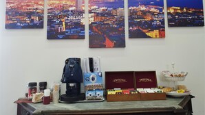 Et bybillede som maleri, en kaffemaskine, Twinings teæsker og en kurv med snacks på et bord.