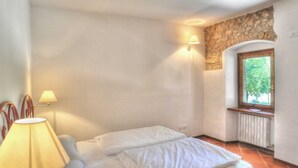 Une chambre à coucher avec un lit, deux lampes fixées au mur, un mur en pierre, une fenêtre et un radiateur.