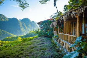 A traditional bamboo structure with a thatched roof, surrounded by lush greenery and mountains in the background.