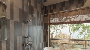 Un baño con una bañera independiente, ducha y un amplio ventanal con vistas a árboles y una estructura de techo de paja.