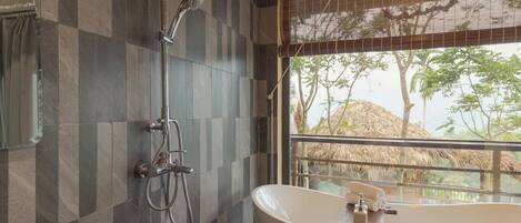 A bathroom with a freestanding bathtub, a shower, and a large window with a view of trees and a thatched roof structure.