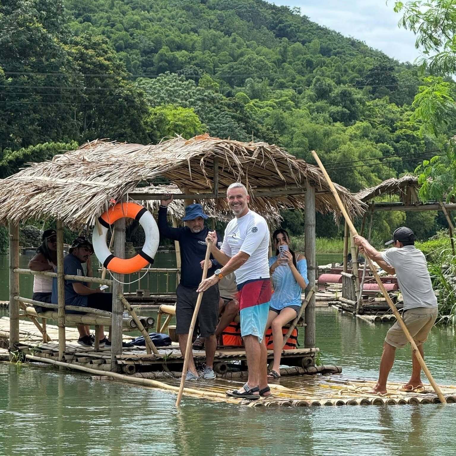 A group of people on a floating structure with a thatched roof, surrounded by water and greenery.