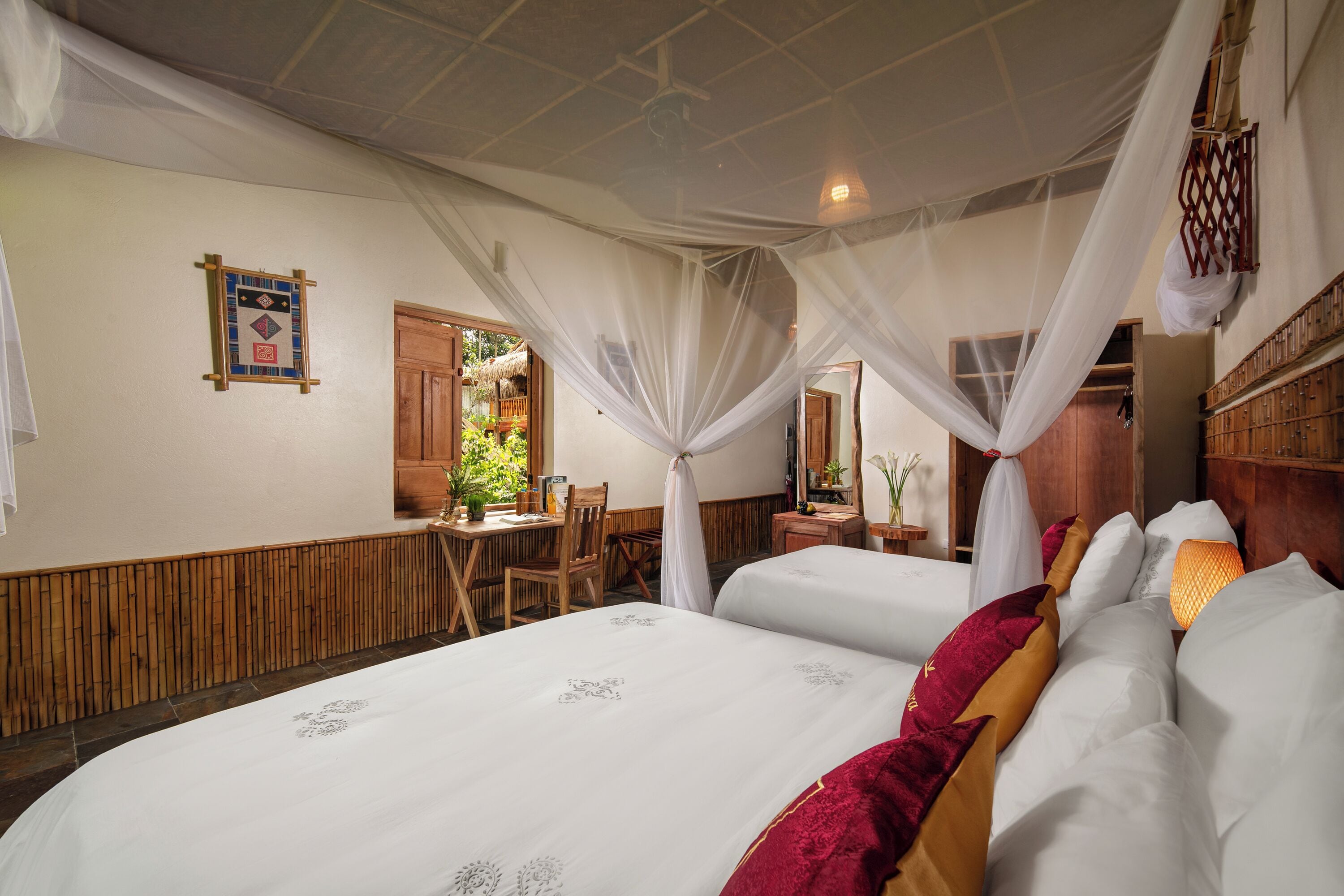 A room with two beds, a wooden table, chairs, and a window with a view of greenery.
