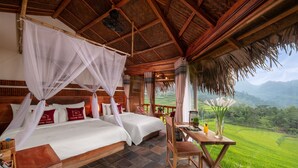 A bedroom with two beds, a wooden table, and a view of green fields. - Puluong Natura Bungalow (Pu Luong)