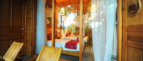 Fachada de madera con puerta de entrada de vidrio, una cama con manta roja y dos sillas de mimbre afuera.