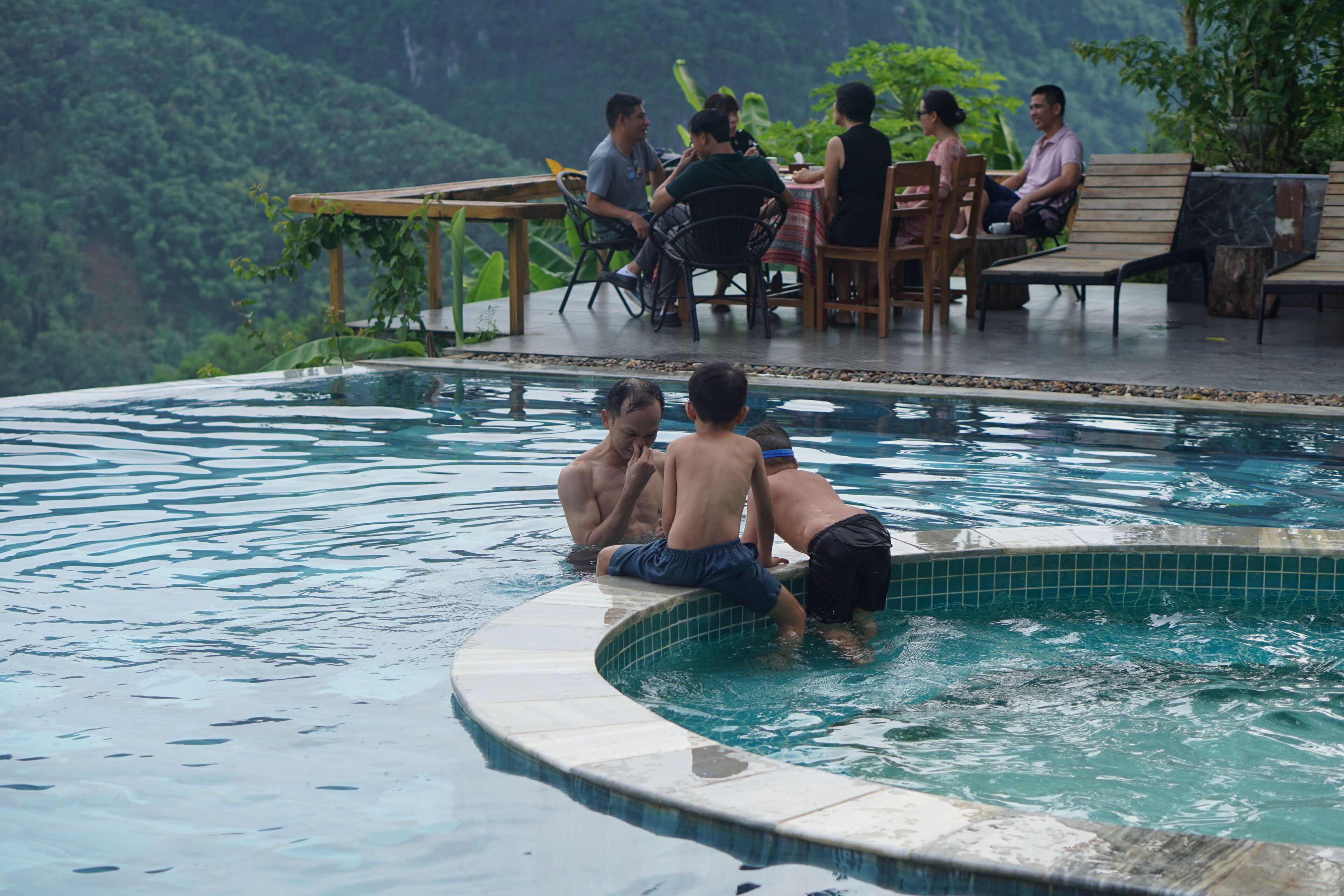 A pool area with a curved edge, a group of people sitting at a table, and a mountainous landscape in the background.