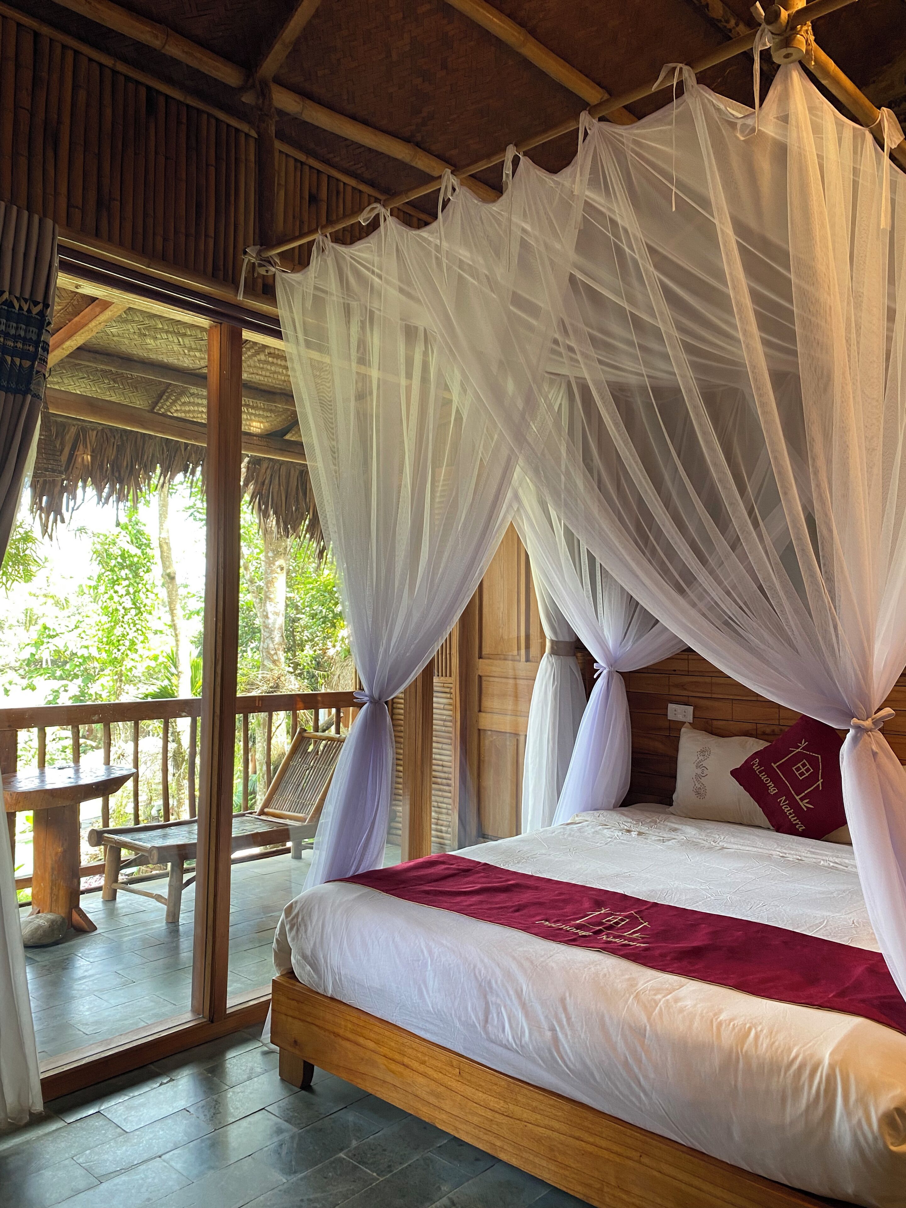 Una habitación con cama, suelo de madera, balcón con mesa y sillas, y vista a la vegetación exterior.