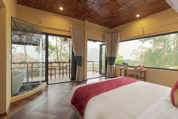 Un dormitorio con una cama grande, un balcón acristalado con un jacuzzi y vistas a árboles y montañas.