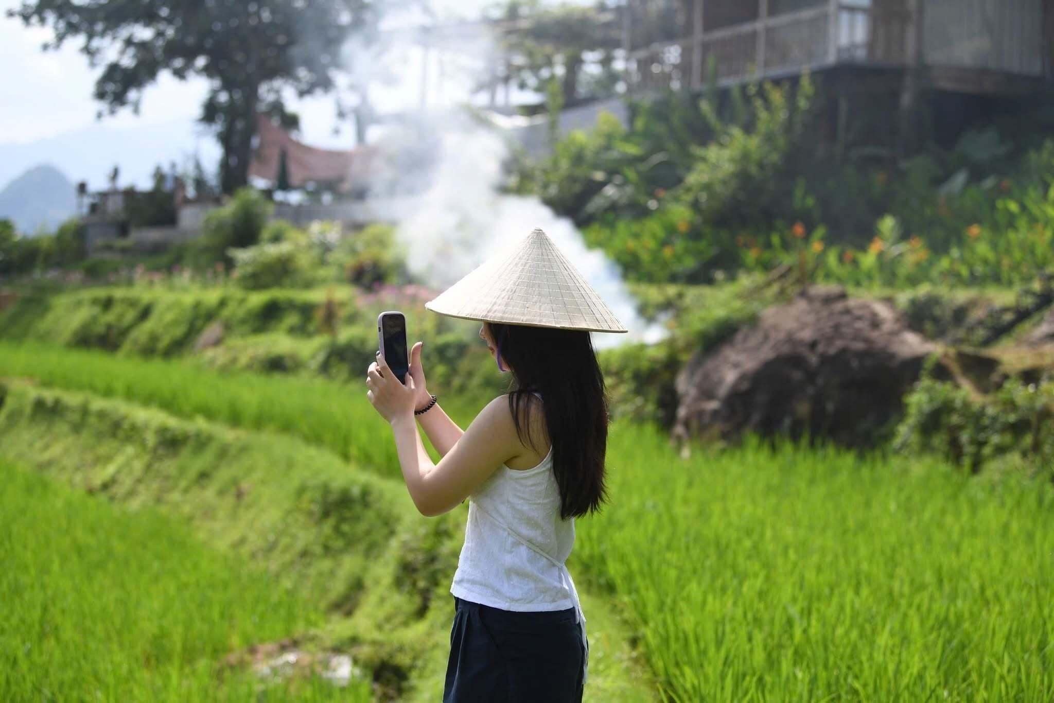 A person taking a photo with a smartphone in a green field with terraced hills in the background.