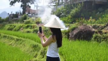 A person taking a photo with a smartphone in a green field with terraced hills in the background.