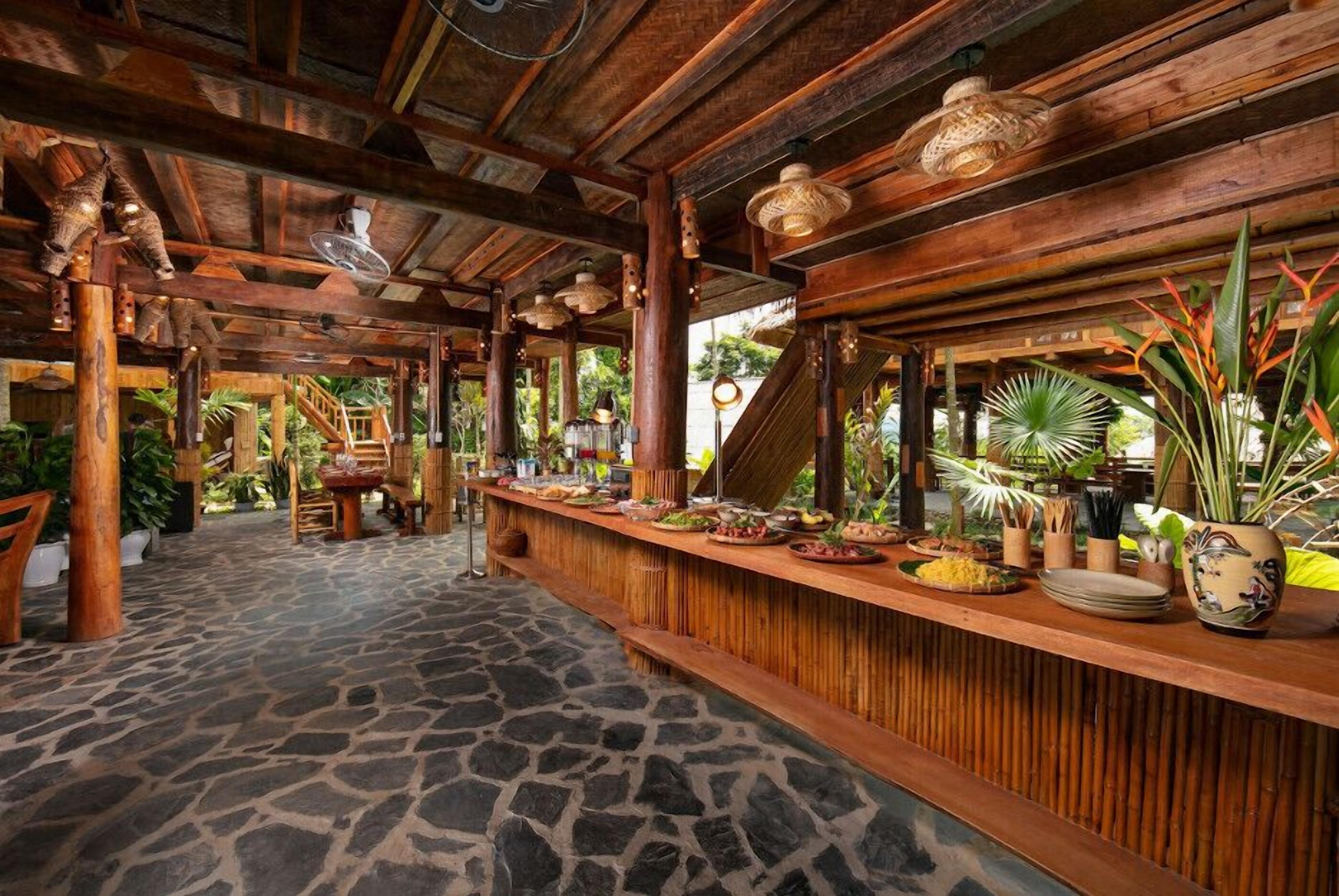 Una zona de bar con barra de madera, suelo de piedra, rodeada de plantas y decoración tropical.