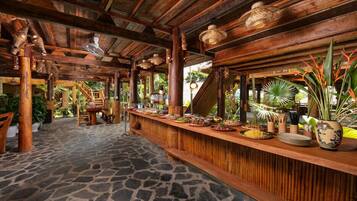 Una zona de bar con barra de madera, suelo de piedra, rodeada de plantas y decoración tropical.