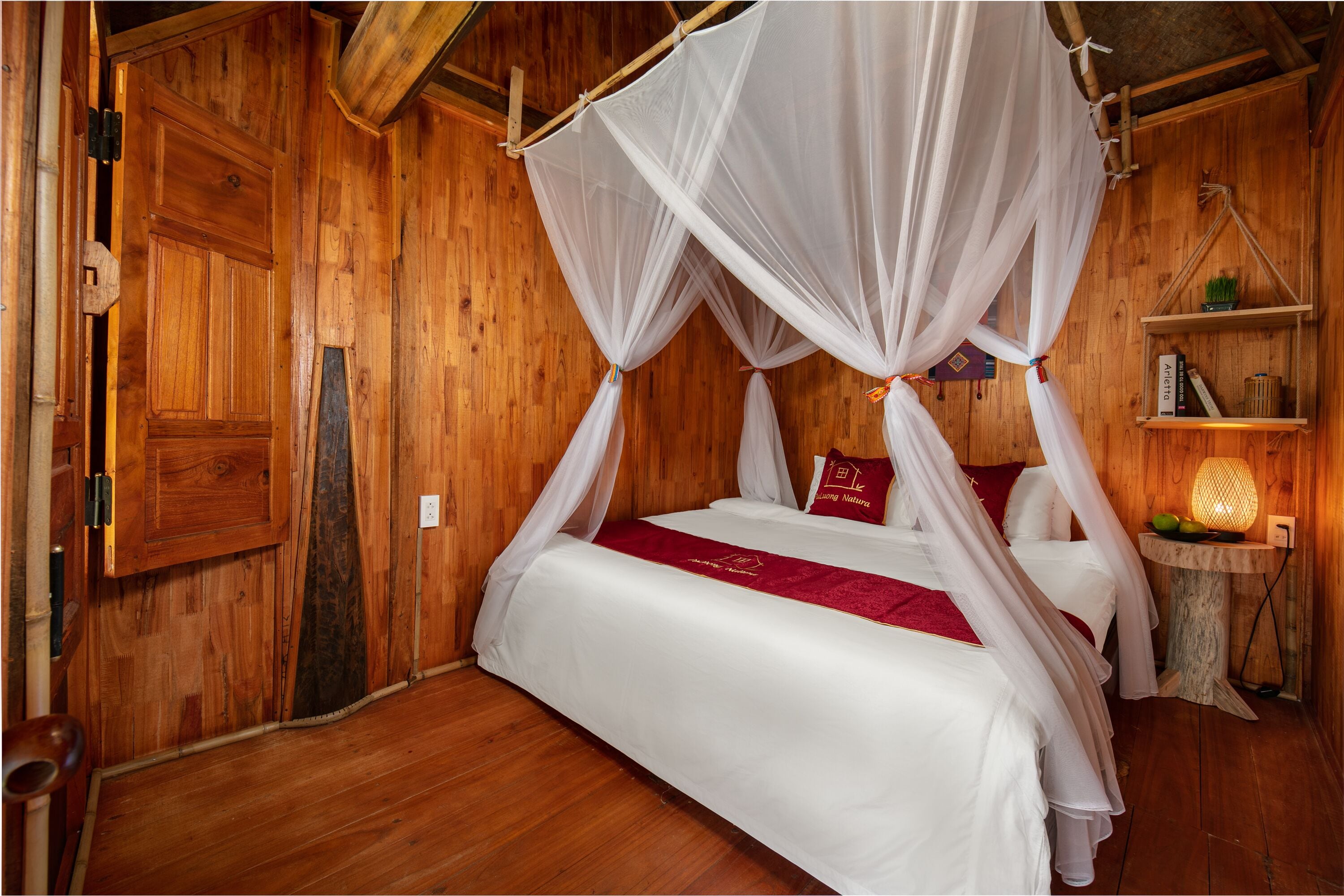 A wooden room with a four-poster bed, a small table with a lamp, and a shelf with books and a plant.