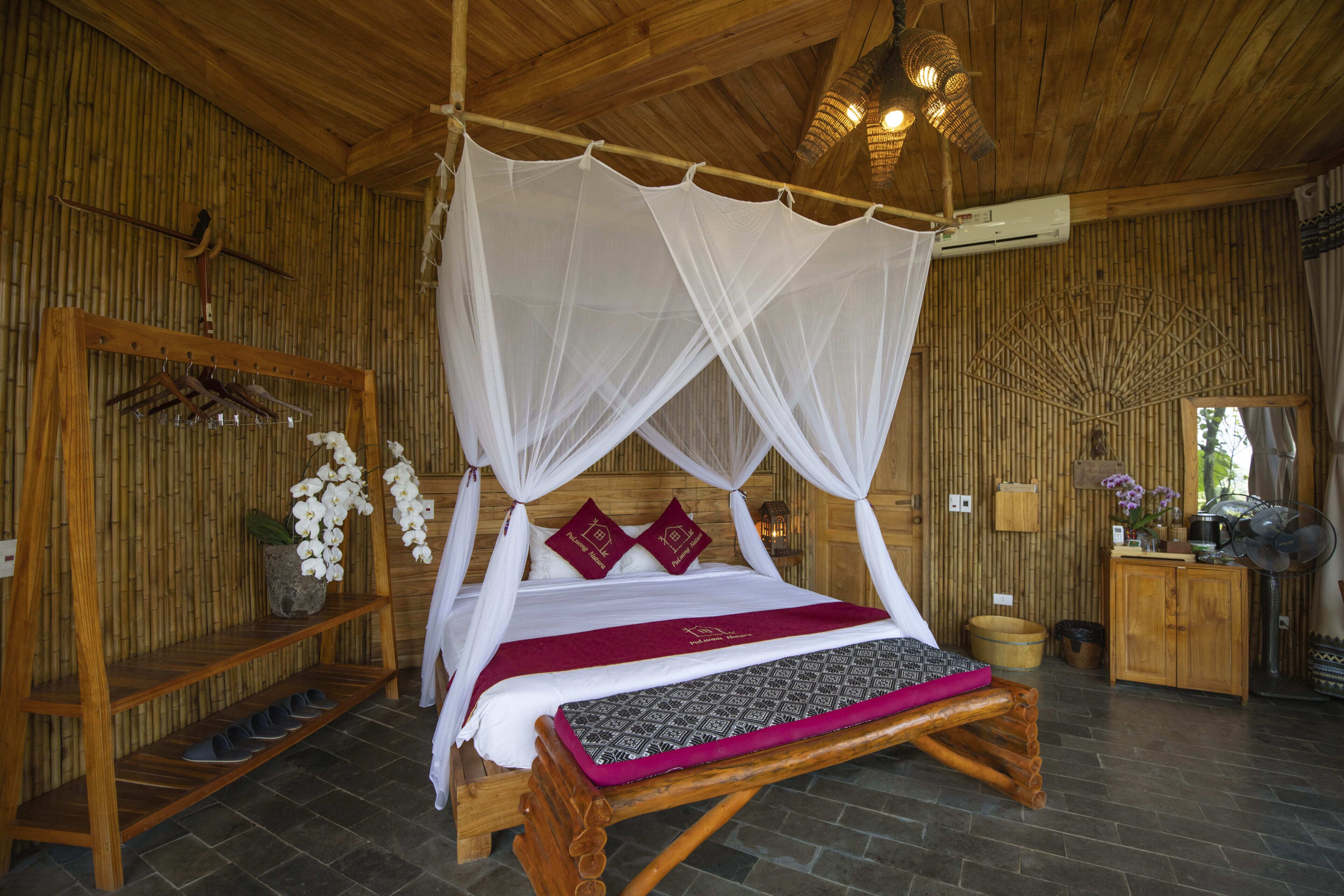 A wooden four-poster bed with a canopy, a shelf with shoes, and a small table with a lamp.
