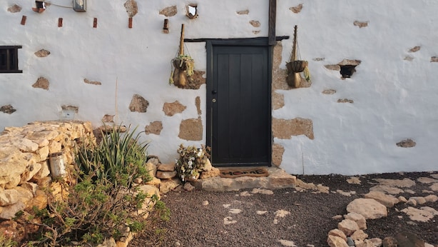 A white building with a black door, stone wall, and a small garden area with rocks and plants. - Villa Cecilio (Puerto del Rosario)