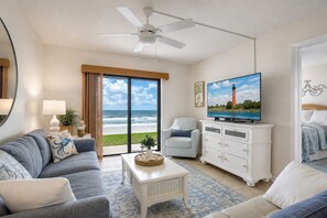 A living room with a large TV, a ceiling fan, a view of the ocean, and a beach chair. - 116 - Oceanfront First-Floor Escape | Steps to the Sand (New Smyrna Beach)