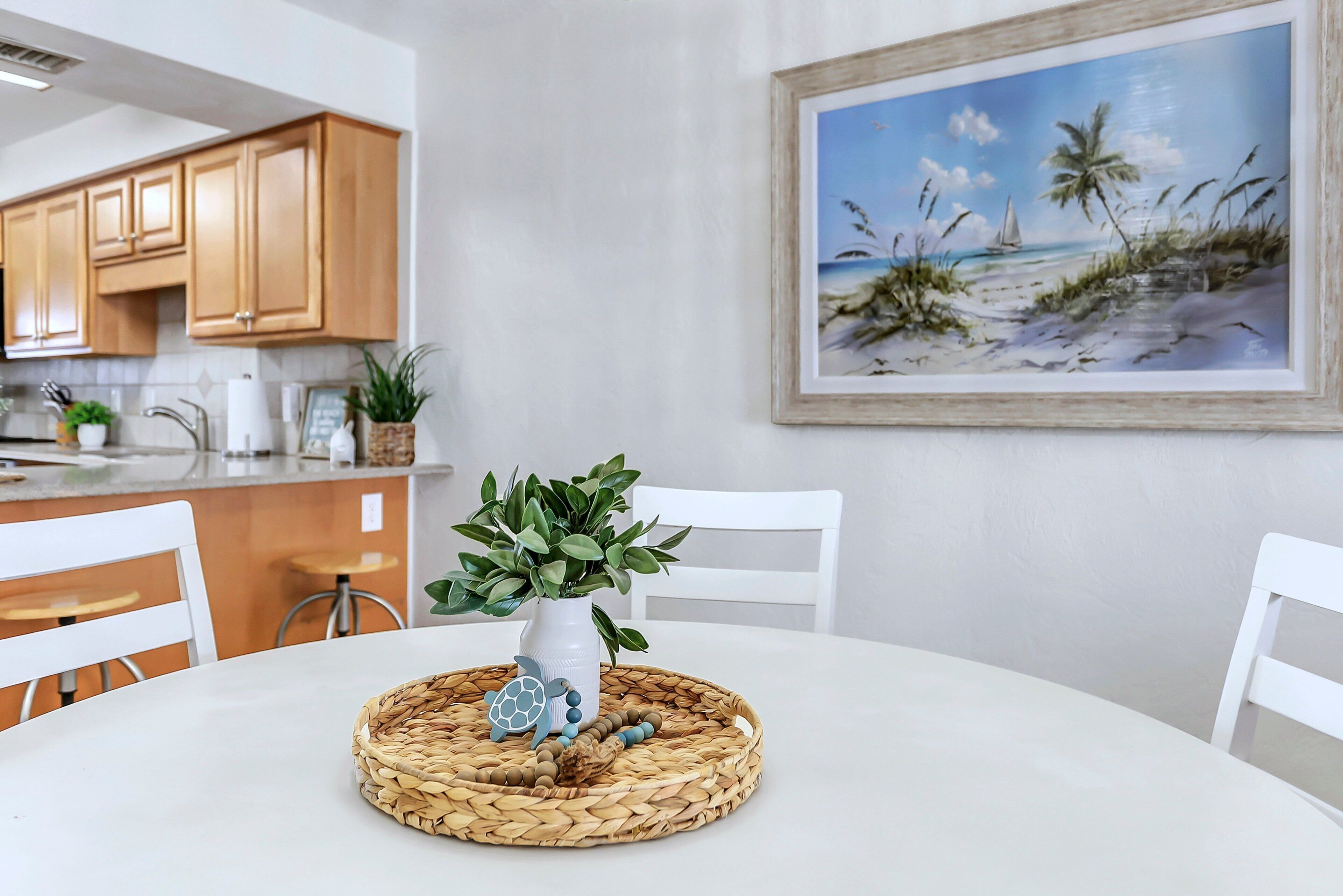 A dining area with a white table, chairs, a woven placemat, a potted plant, and a framed beach painting on the wall.