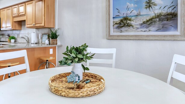 Ein Essbereich mit einem weißen Tisch, Stühlen, einer gewebten Tischmatte, einem Blumentopf und einem gerahmten Strandbild an der Wand.