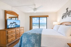 A hotel room with a bed, a television on a wooden dresser, a ceiling fan, and a view of the ocean through large windows. - 116 - Oceanfront First-Floor Escape | Steps to the Sand (New Smyrna Beach)