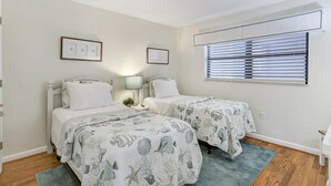 A twin bedroom with two beds, a window with blinds, a nightstand with a lamp, and framed pictures on the wall.