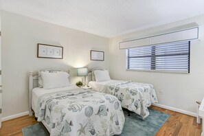 A twin bedroom with two beds, a window with blinds, a nightstand with a lamp, and framed pictures on the wall. - 116 - Oceanfront Delight Ground Floor (New Smyrna Beach)