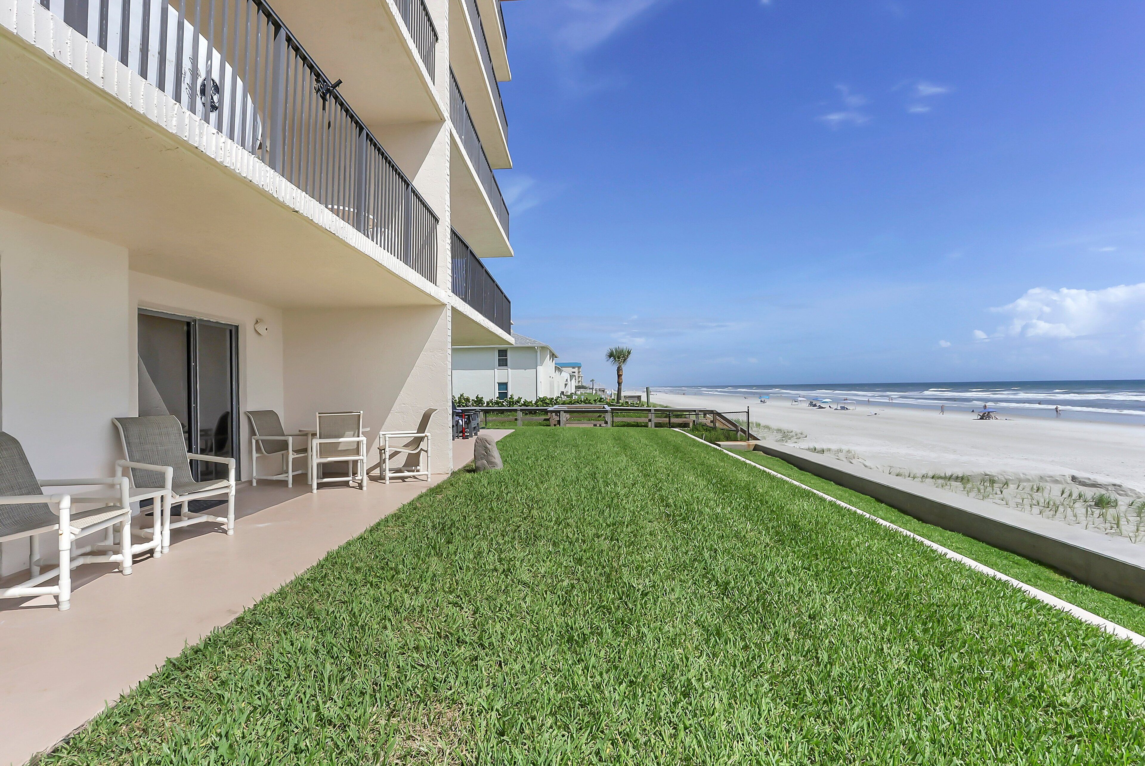 A hotel balcony with a view of the beach, outdoor seating, and a well-maintained lawn.