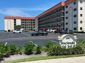 A multi-story hotel with a parking lot, a sign indicating the address, and a view of the ocean. - 116 - Oceanfront Delight Ground Floor (New Smyrna Beach)