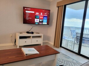 Una habitación con televisión de pantalla plana montada en la pared, un mueble blanco debajo, una mesa de madera con un libro, y un balcón con vista al océano.