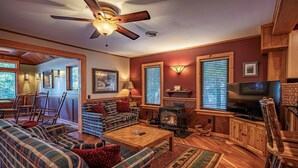 A cozy living room with plaid furniture, a fireplace, a television, and wooden accents. - Historic Highland Cottage - 4th Lake (Inlet)