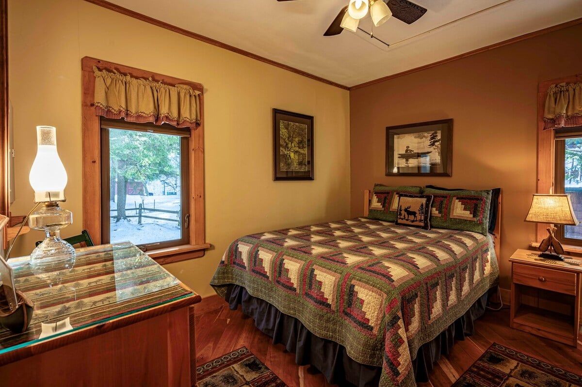 A bedroom with a large bed, two windows with curtains, a ceiling fan, and a bedside table with a lamp.