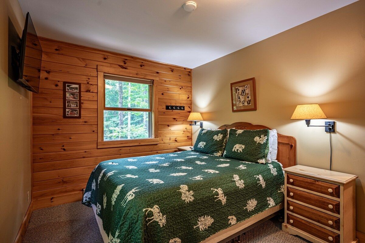 Une chambre avec un mur lambrissé en bois, un lit recouvert d’une couette verte, deux lampes de chevet et une fenêtre donnant sur des arbres.