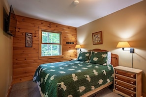 Une chambre avec un mur lambrissé en bois, un lit recouvert d’une couette verte, deux lampes de chevet et une fenêtre donnant sur des arbres.
