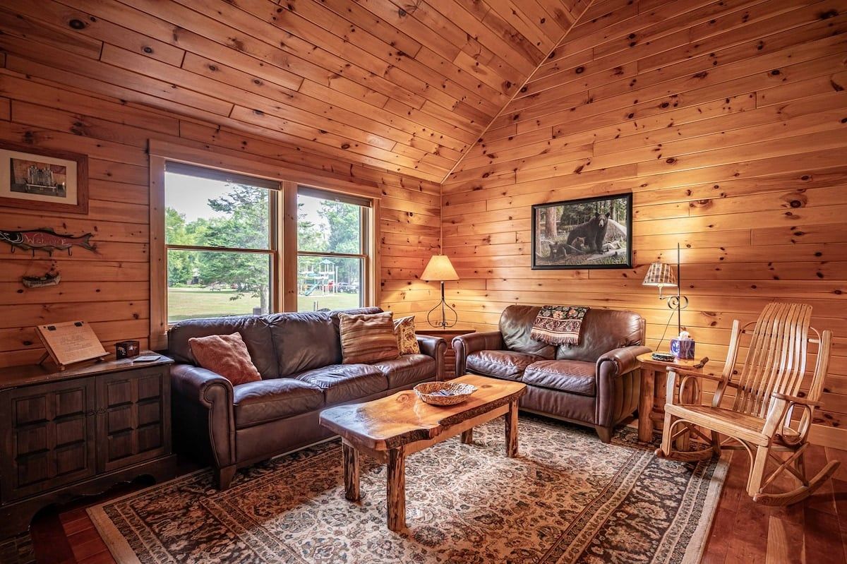 Ein Interieur einer Holzhütte mit Holzbalkendecke, Ledersofas, einem Couchtisch aus Holz und einem Schaukelstuhl.