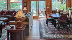 L’intérieur chaleureux d’un chalet avec un plafond en bois, un canapé, une table à manger avec des chaises et une vue sur un lac, visible à travers de grandes fenêtres.