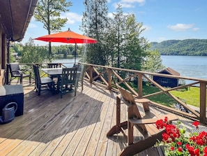 A wooden deck with outdoor furniture, a table, and chairs, overlooking a lake with a cabin in the background. - Chalet on 4th - Lakefront Vacation Rental (Old Forge)