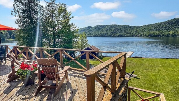Une terrasse en bois, avec des chaises et une table, donnant sur un lac, avec une colline boisée en arrière-plan.