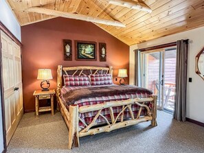 A rustic bedroom with a wooden bed frame, a red plaid bedspread, two bedside lamps, and a large window with curtains.