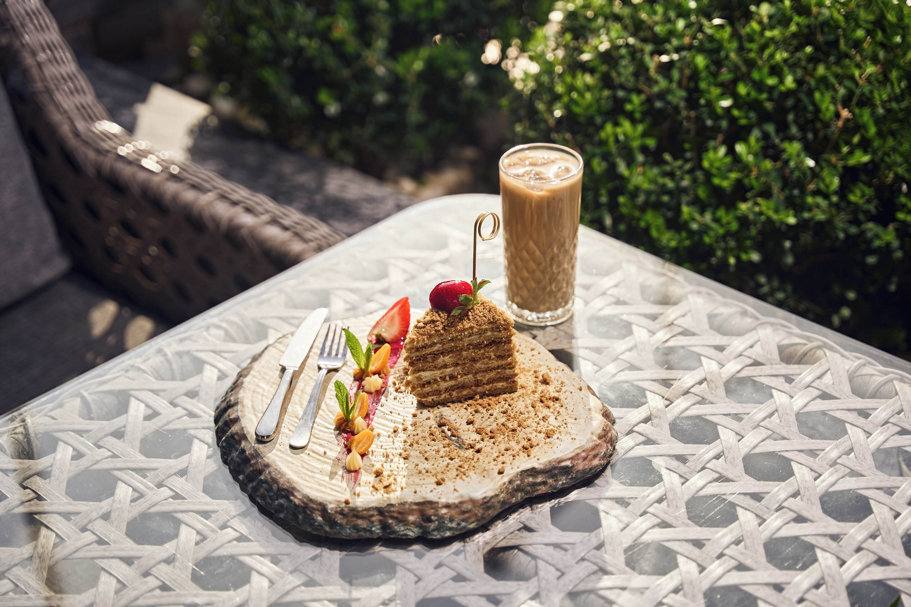 Un postre sobre una mesa de vidrio con textura tejida, acompañado por un tenedor, un cuchillo y un vaso de bebida helada.