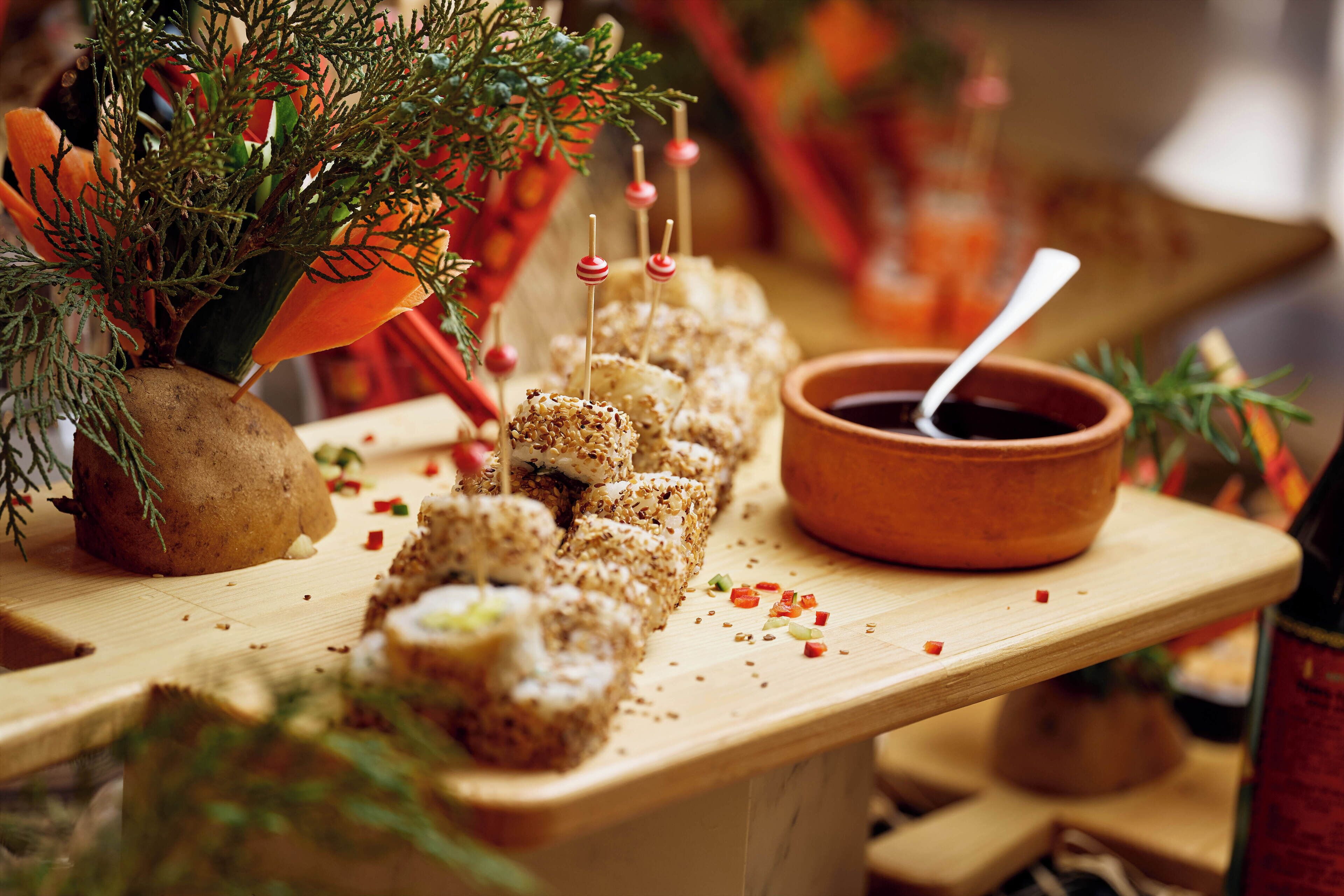 A wooden serving board with skewered food items, a small bowl of dipping sauce, and decorative greenery.