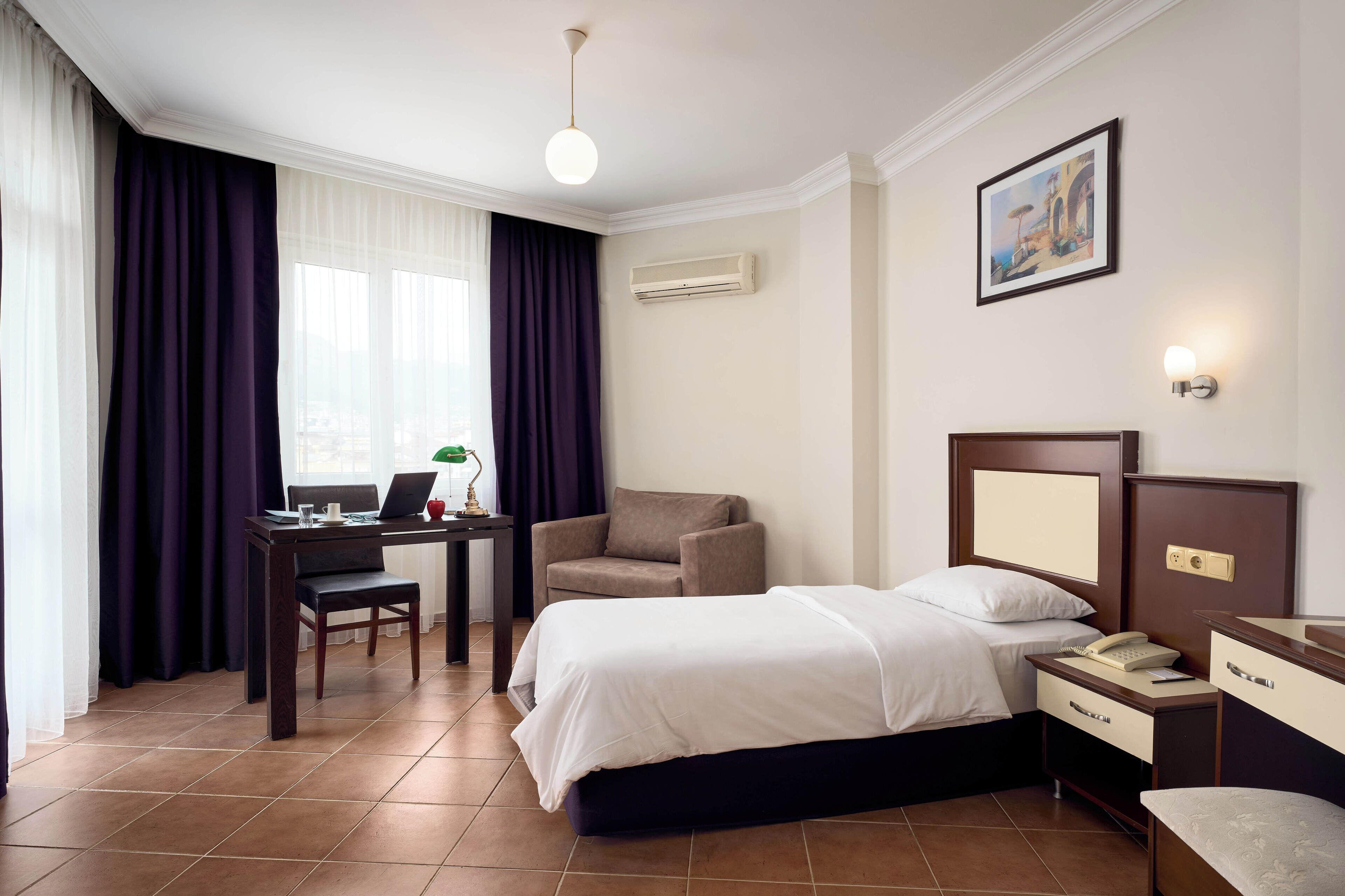 A hotel room with a bed, desk, chair, and a painting on the wall.