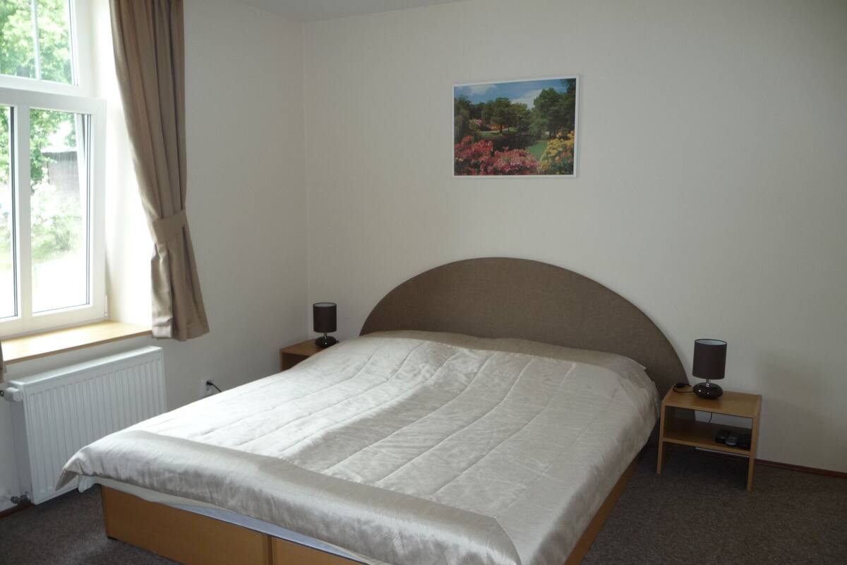 A bedroom with a bed, two bedside tables, a window with curtains, a radiator, and a painting on the wall.