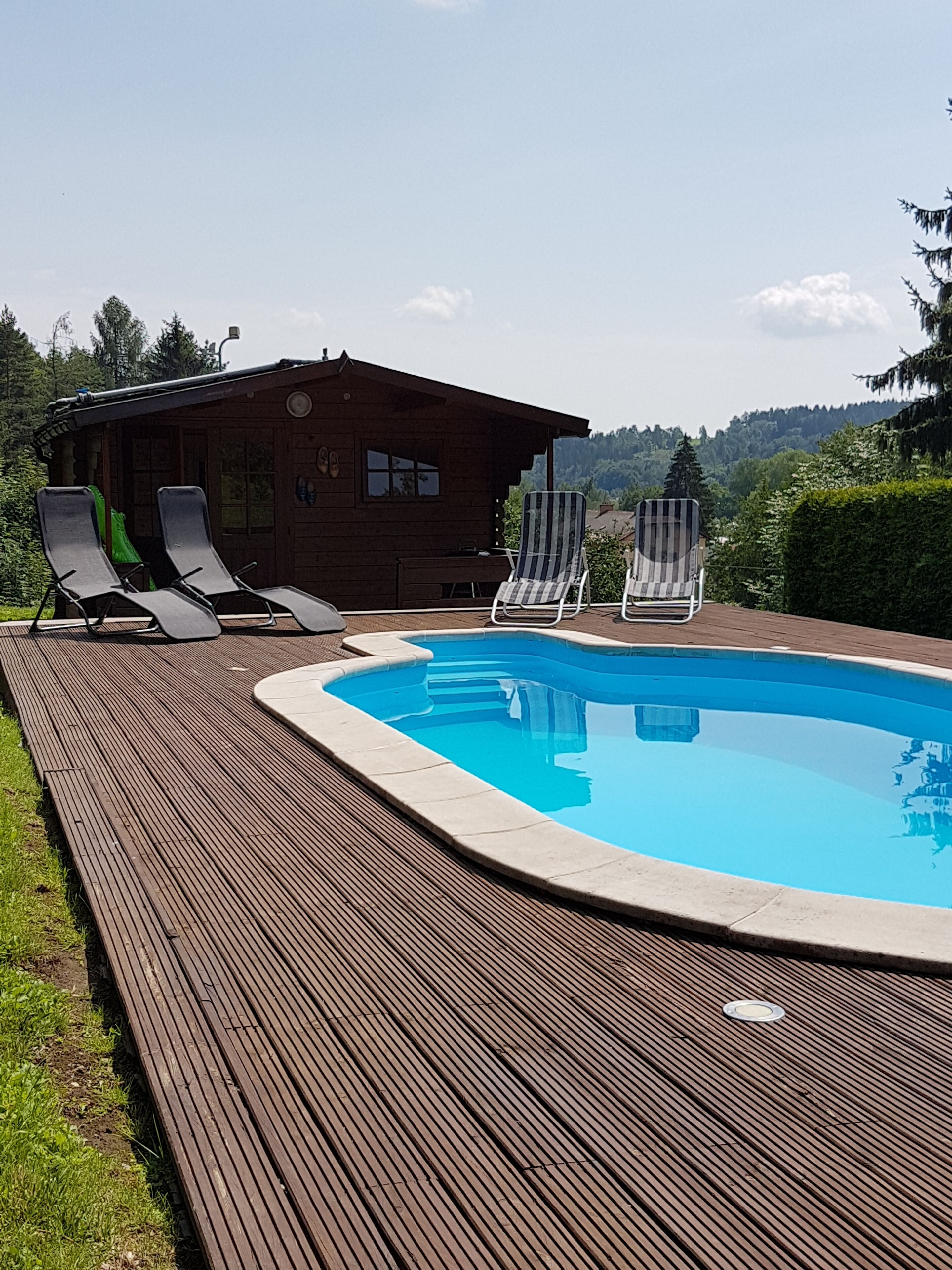 A wooden deck with a swimming pool, two lounge chairs, and a wooden cabin in the background.
