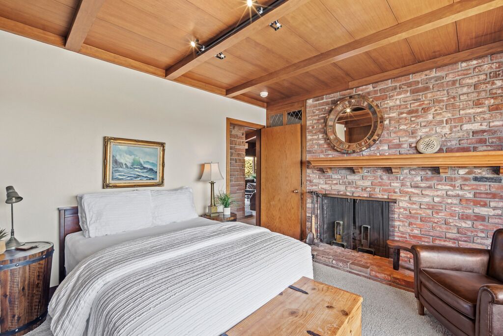 Une chambre avec un grand lit, un plafond en bois, une cheminée en briques et un fauteuil en cuir.