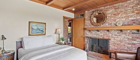 Une chambre avec un grand lit, un plafond en bois, une cheminée en briques et un fauteuil en cuir.