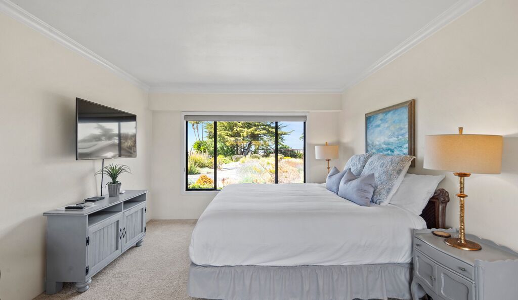 A bedroom with a large bed, a TV mounted on the wall, a window with a view of trees, and a bedside table with a lamp.