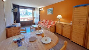 A room with a dining table set for four, a sofa, a wooden cabinet, and a large window with a view. - Studio for 4 at slopes, center Les Deux Alpes (Les Deux Alpes)