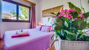 Un dormitorio con una cama, una mesita de noche con un objeto rosa y una planta en maceta con flores rosadas.