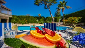Una mesa junto a la piscina con sandía, naranjas, cerezas y vasos de agua y una bebida.
