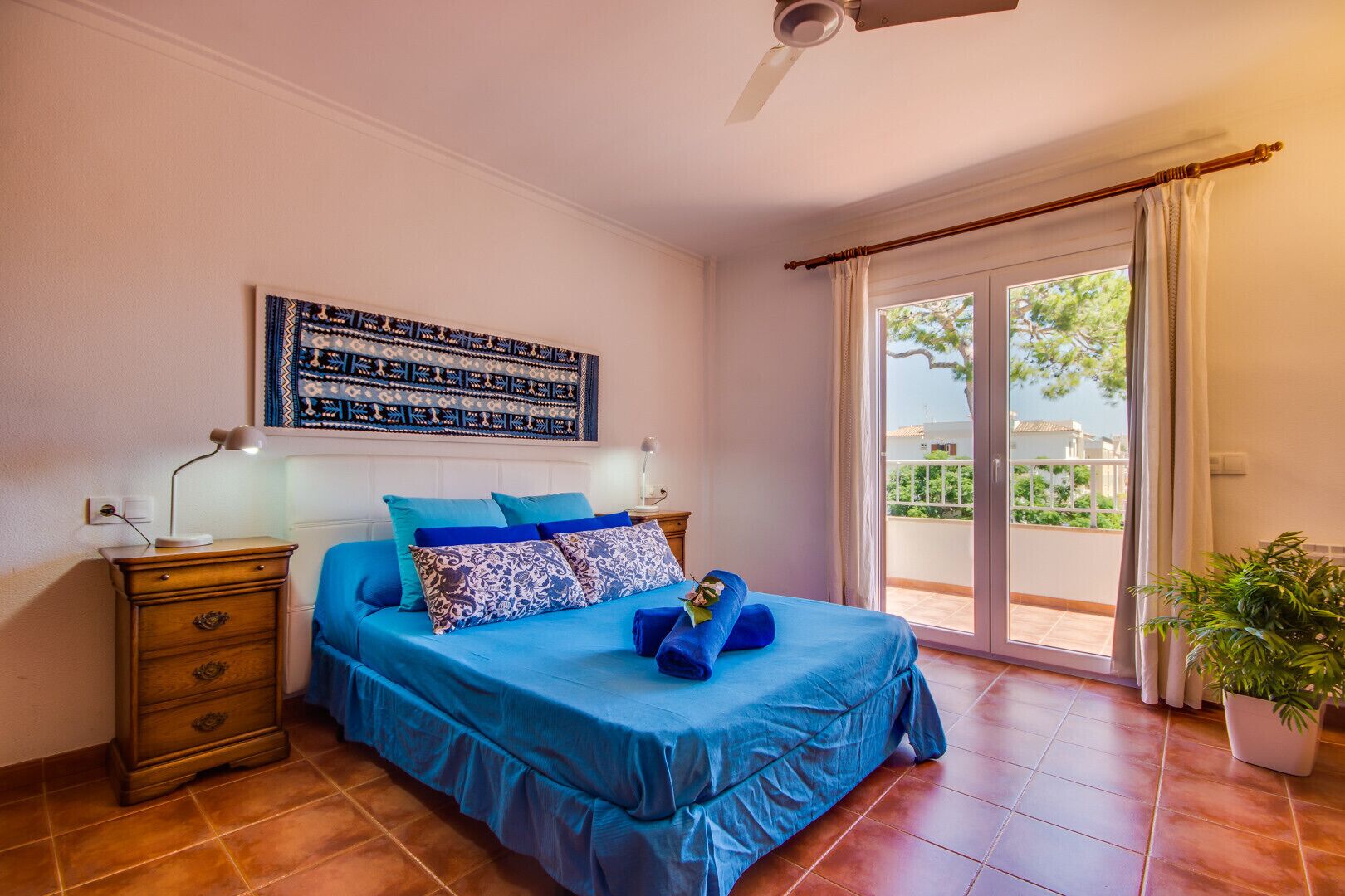 Une chambre à coucher comprenant un lit, une table de chevet, un ventilateur de plafond, une fenêtre avec des rideaux et une plante en pot.